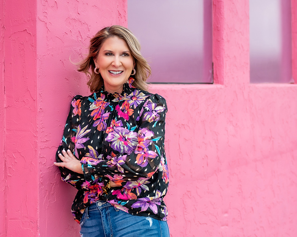 Oklahoma City branding photos of a smiling woman in floral blouse leans against a vibrant pink wall, evoking a cheerful and lively atmosphere. Her arms are crossed casually.