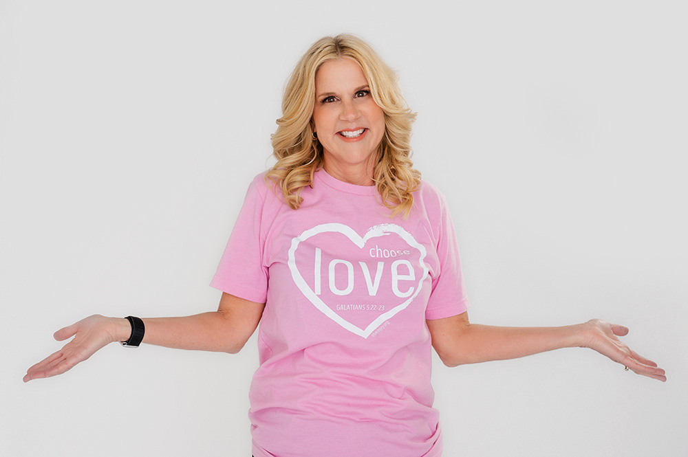 A smiling woman in a pink "choose love" T-shirt poses with open arms against a plain background. Her expression is friendly and inviting.