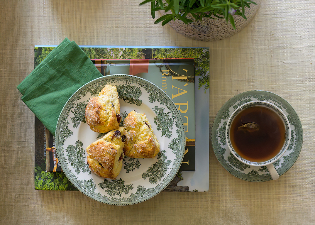 A top view of a table setting with scones on a floral-patterned plate, a green napkin, a cup of tea, and a potted plant beside books. Cozy and inviting.