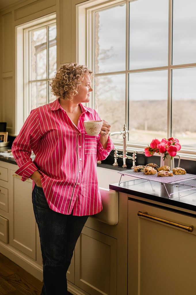 A person with curly hair in a pink striped shirt and jeans gazes out a window while holding a mug. Nearby are cooling cookies and vibrant pink flowers, creating a cozy and warm atmosphere.