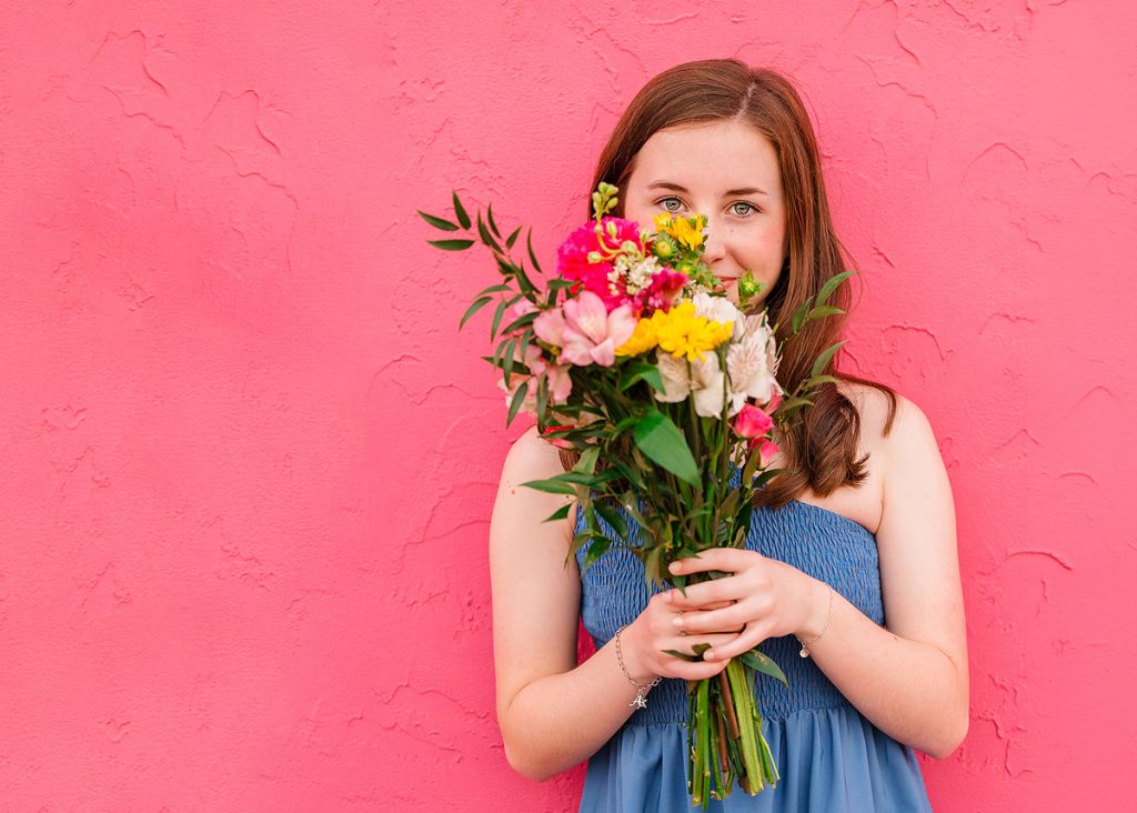 High school senior girl holding flowers in front of pink wall during Oklahoma City senior portrait session