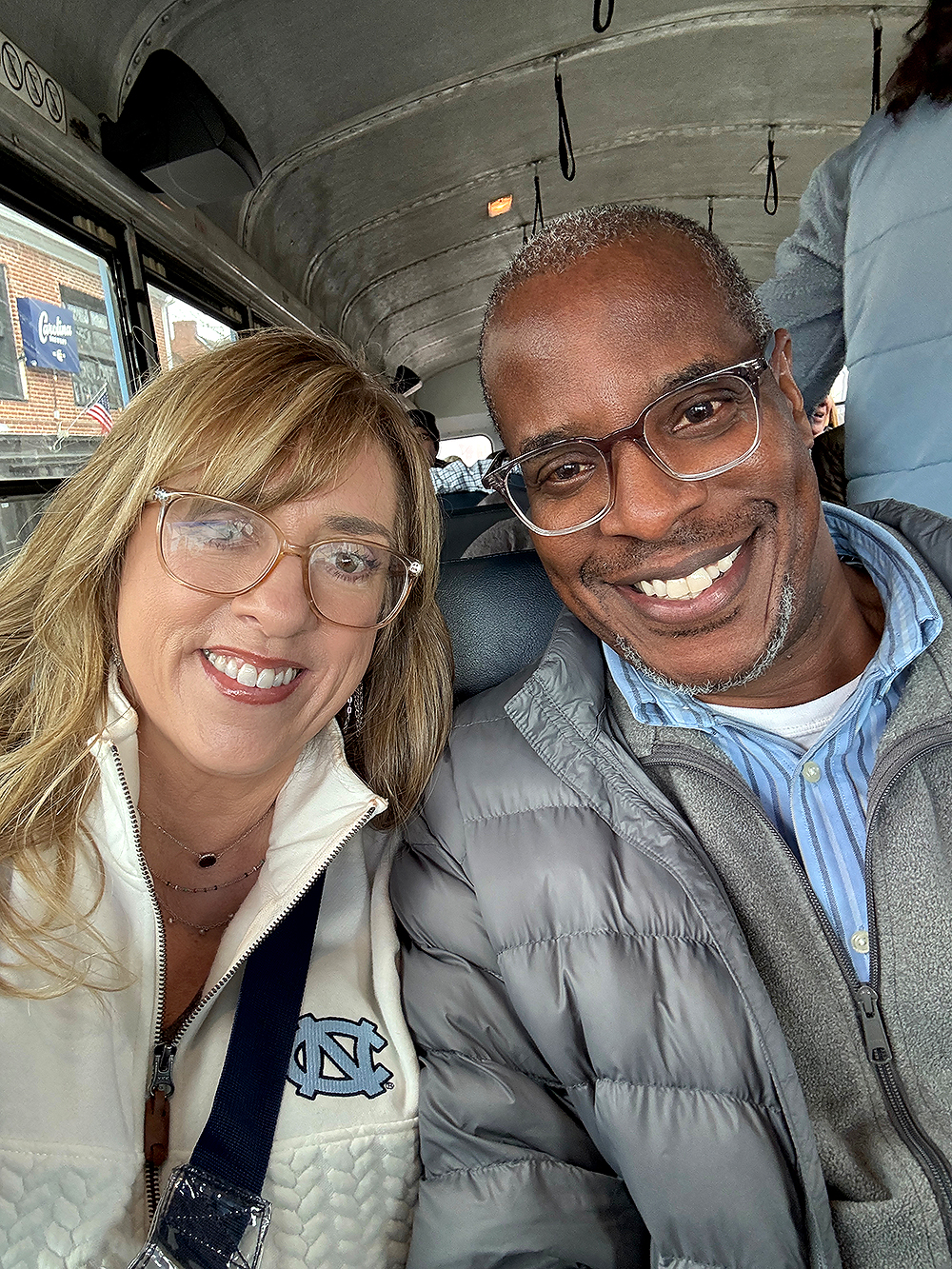 A smiling woman and man sit together on a bus. She wears glasses and a white jacket with a logo. He wears glasses and a gray puffy jacket. The mood is cheerful.