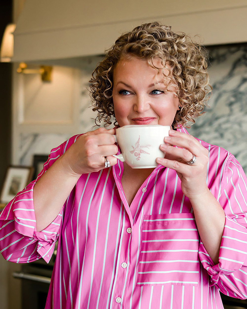A smiling person with curly hair sips from a mug in a cozy kitchen, wearing a pink striped shirt