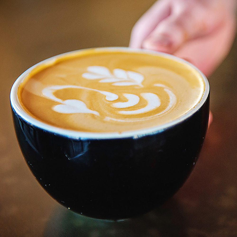 A hand holds a black cup filled with latte art depicting a leafy pattern. 