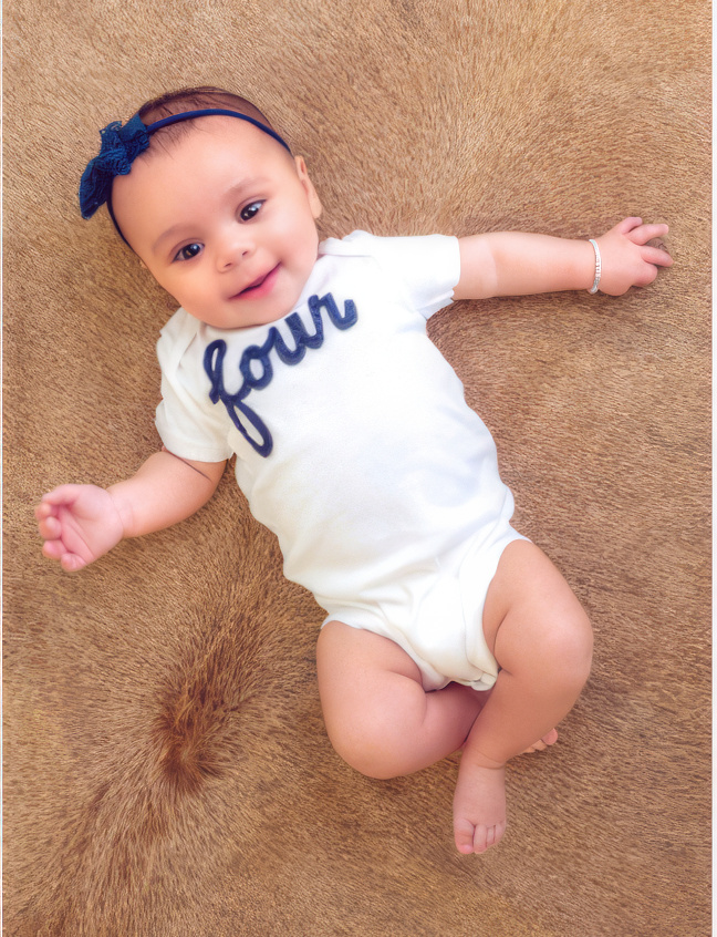 A smiling baby wearing a white onesie with the word "four" lies on a brown, cowhide. The baby has a blue headband and a bracelet.