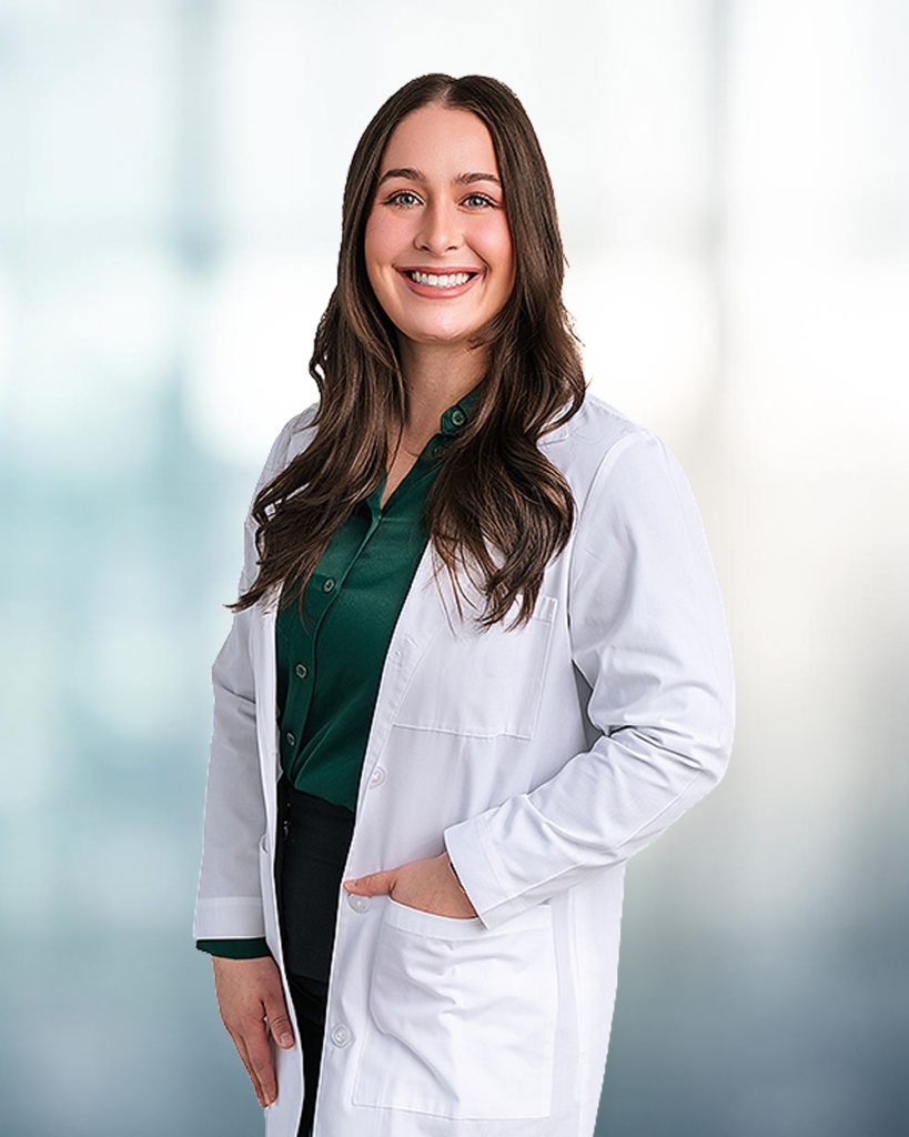 Smiling woman in a white lab coat over a green shirt, standing with hands in pockets.