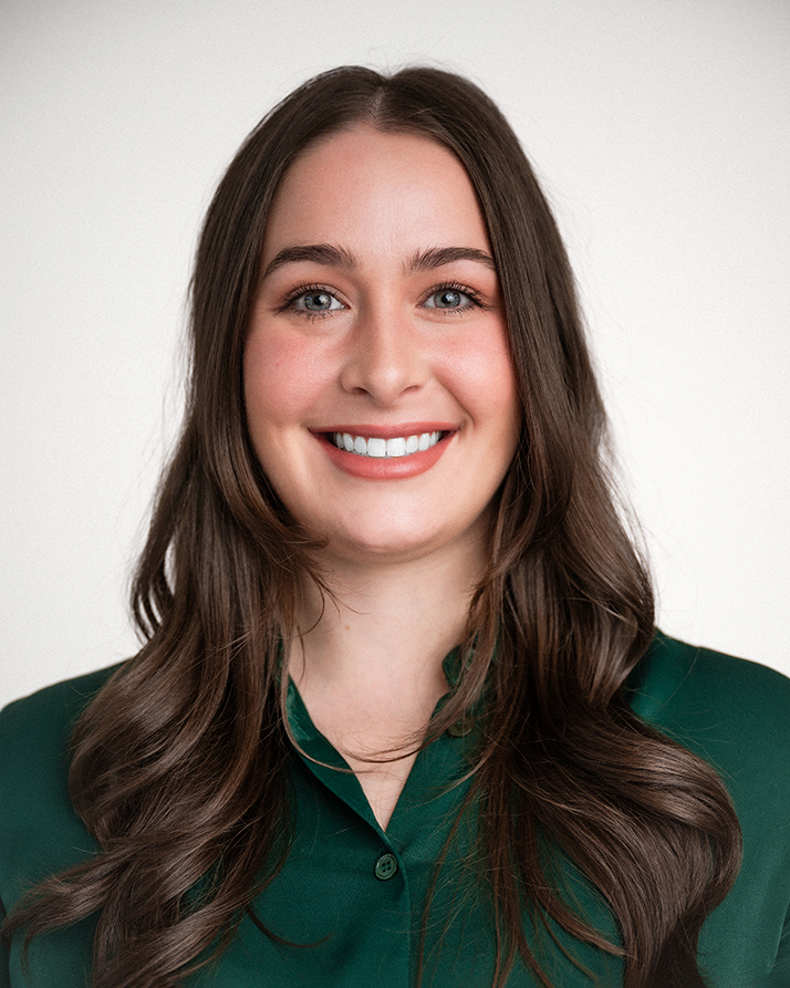 Smiling woman with long brown hair and a bright, welcoming expression, wearing a green blouse. The background is plain white, conveying a professional yet friendly tone.