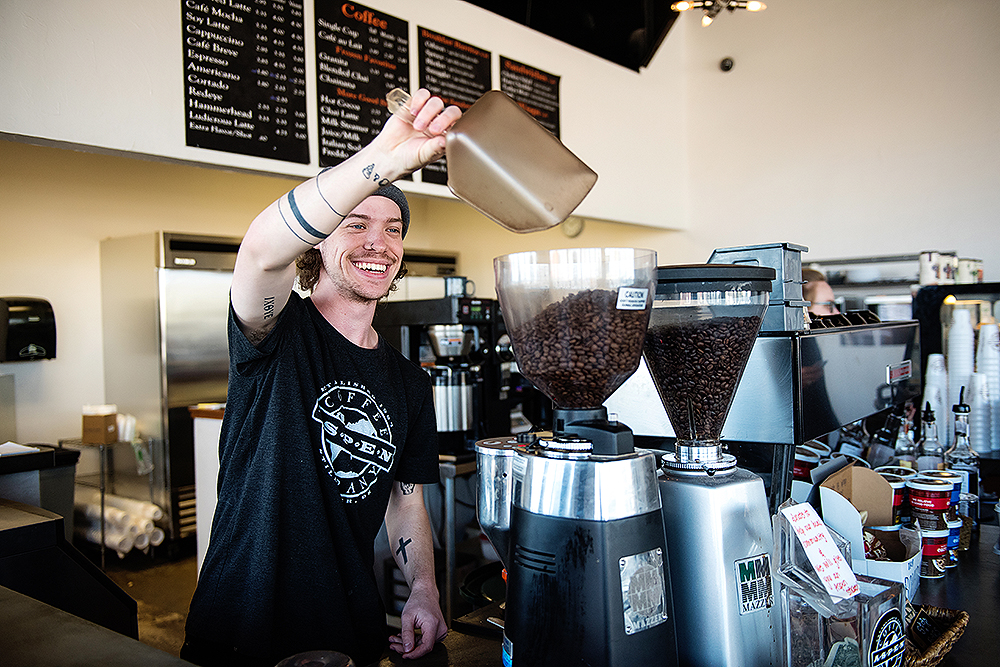 A cheerful barista in a beanie and casual t-shirt pours coffee beans into a grinder at a bright cafe. A menu board is visible in the background.