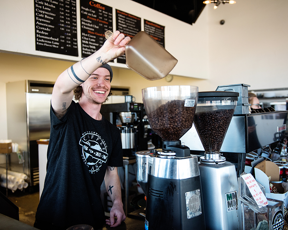 A cheerful barista in a beanie and casual t-shirt pours coffee beans into a grinder at a bright cafe. A menu board is visible in the background.