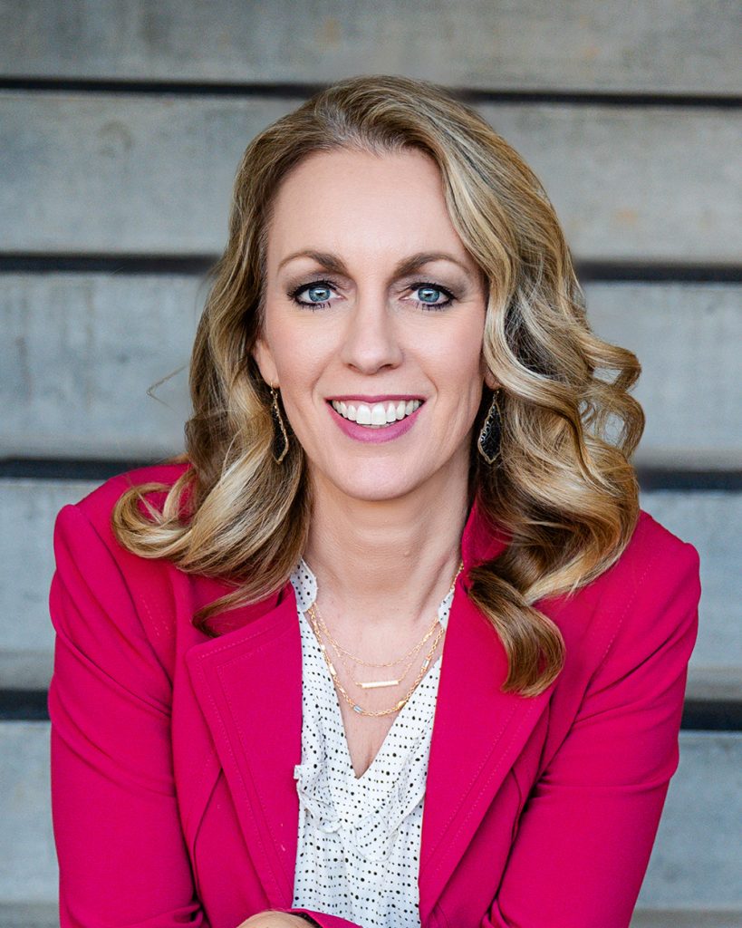 A woman with wavy blond hair and a bright smile wears a vibrant pink blazer and white blouse