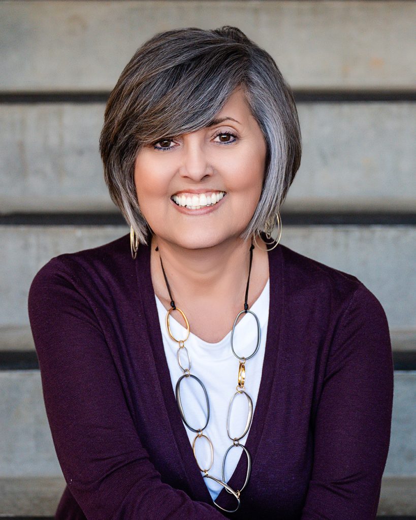 Smiling woman with short gray hair sits on steps. She wears a dark cardigan, a chunky necklace, and exudes warmth and confidence.