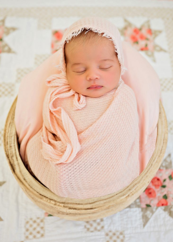 A newborn baby peacefully sleeps, wrapped in a soft peach knit blanket with a matching bonnet, nestled in a cozy basket on a floral quilt.