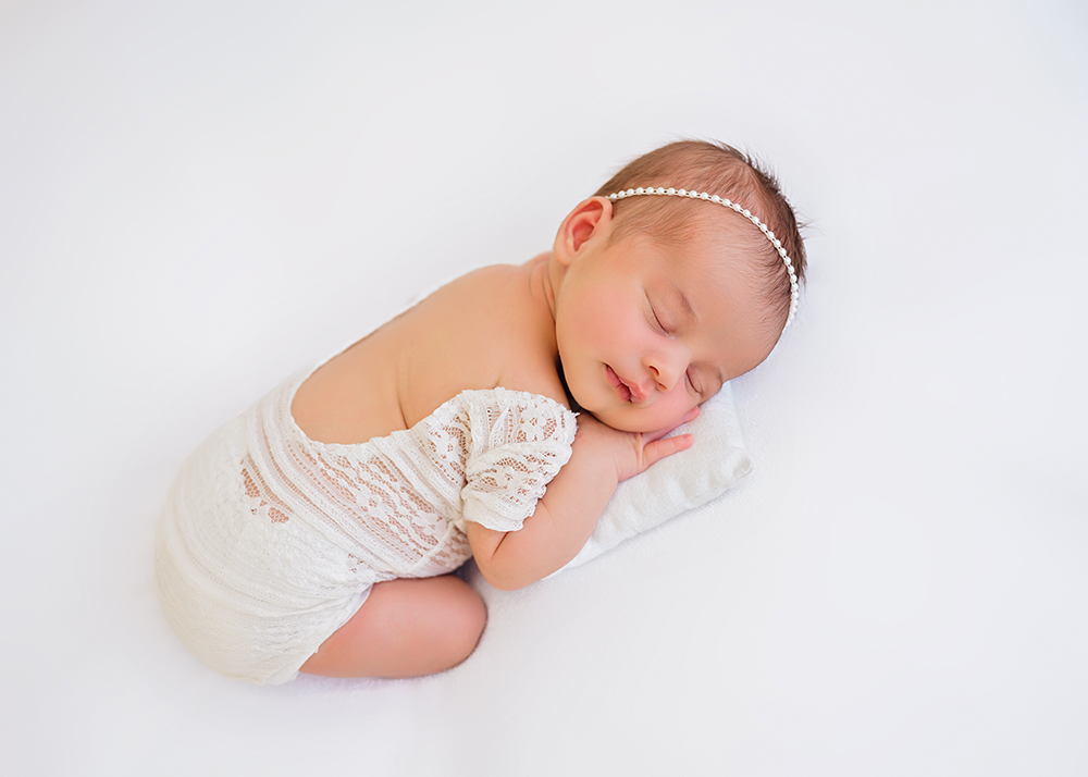 A newborn baby peacefully sleeps on its side, wearing a delicate white lace outfit and a pearl headband, on a soft white background.