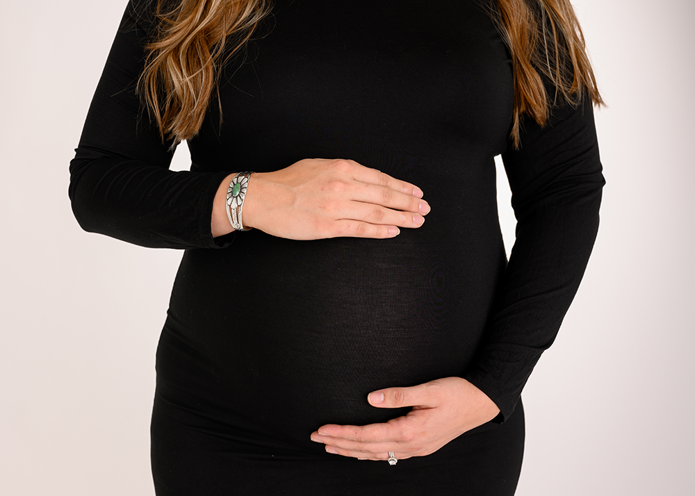 A pregnant woman in a black dress cradles her belly with both hands.