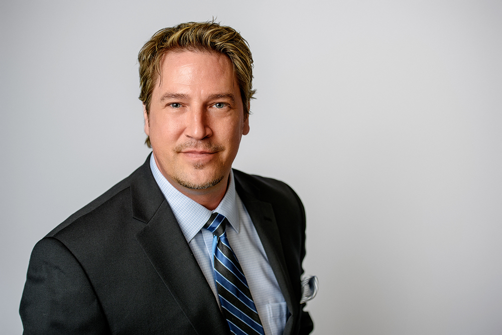 A man in a suit and striped tie smiles slightly against a plain background