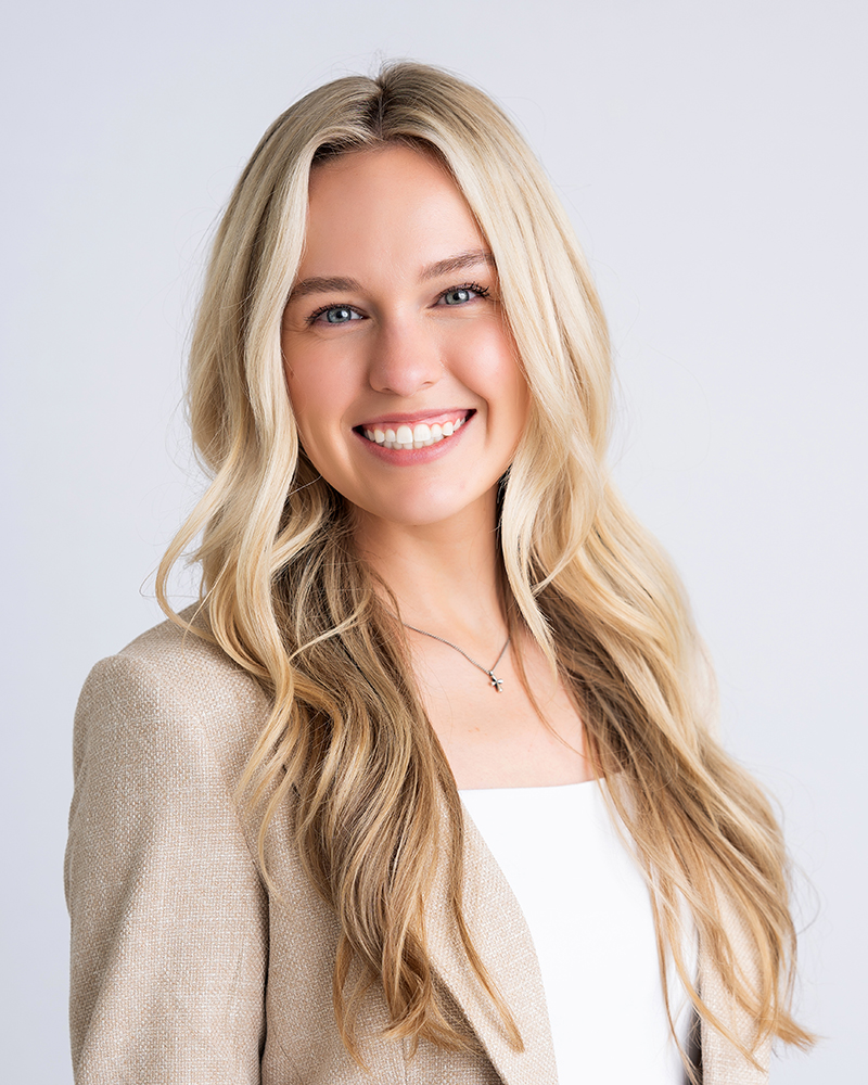 A smiling woman with long blonde hair wears a beige blazer and white top against a plain background. She conveys a professional and friendly demeanor.