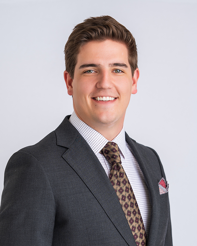 Young man in a dark suit, white checkered shirt, and patterned tie, smiling confidently against a plain light background, conveying professionalism.