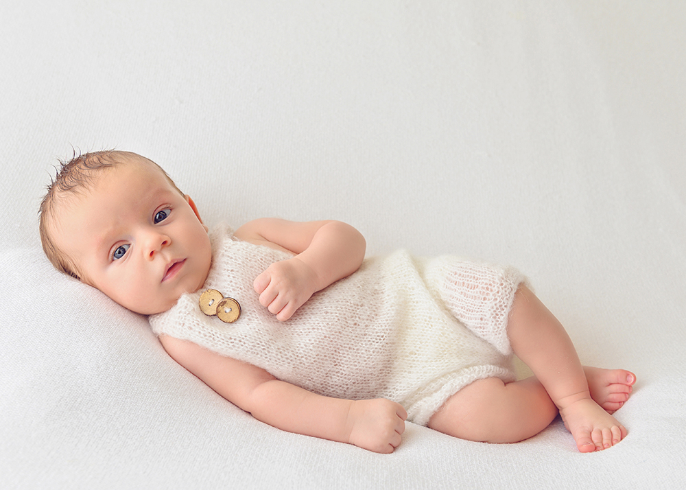 A baby lies on a soft white blanket, wearing a knitted cream romper with wooden buttons
