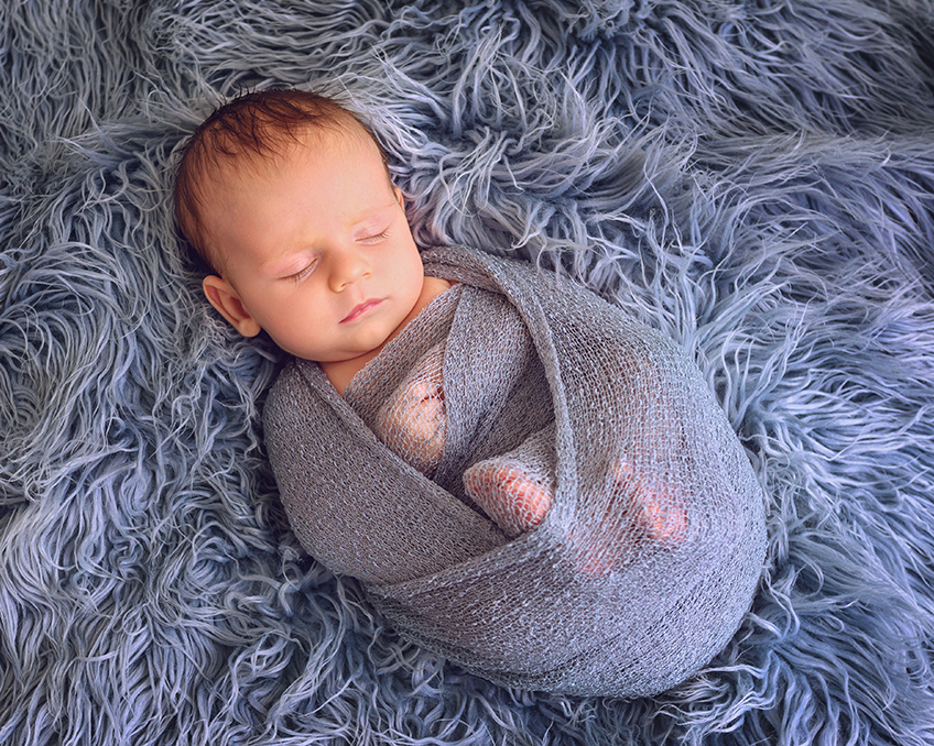 A sleeping baby wrapped in a gray blanket rests on a fluffy, textured blue surface.