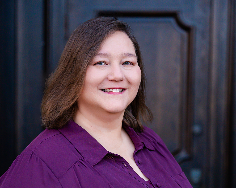 Smiling woman with shoulder-length brown hair wearing a purple shirt stands in front of a dark wooden door, conveying a warm and friendly demeanor