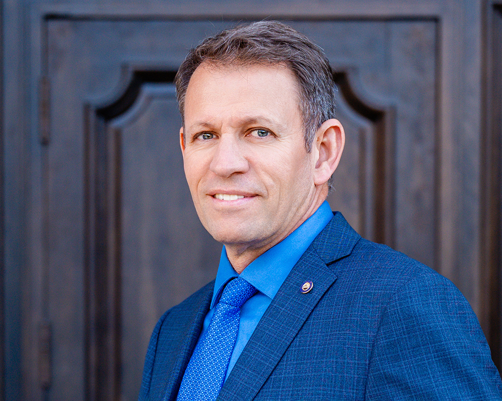A realtor in a blue suit and tie stands in front of a dark wooden door. He has light hair, a confident expression, and a pin on his lapel.