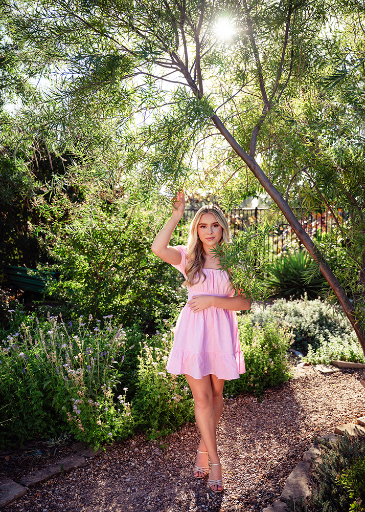 A high school senior poses in a light pink dress on a garden path, surrounded by lush greenery and soft sunlight, conveying a serene and peaceful mood.