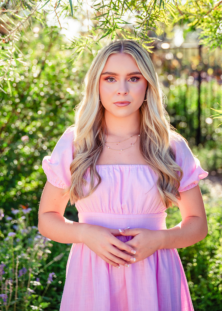 High school senior with long blonde hair, wearing a pink dress, stands in a sunlit garden. Her expression is serene, surrounded by lush greenery.