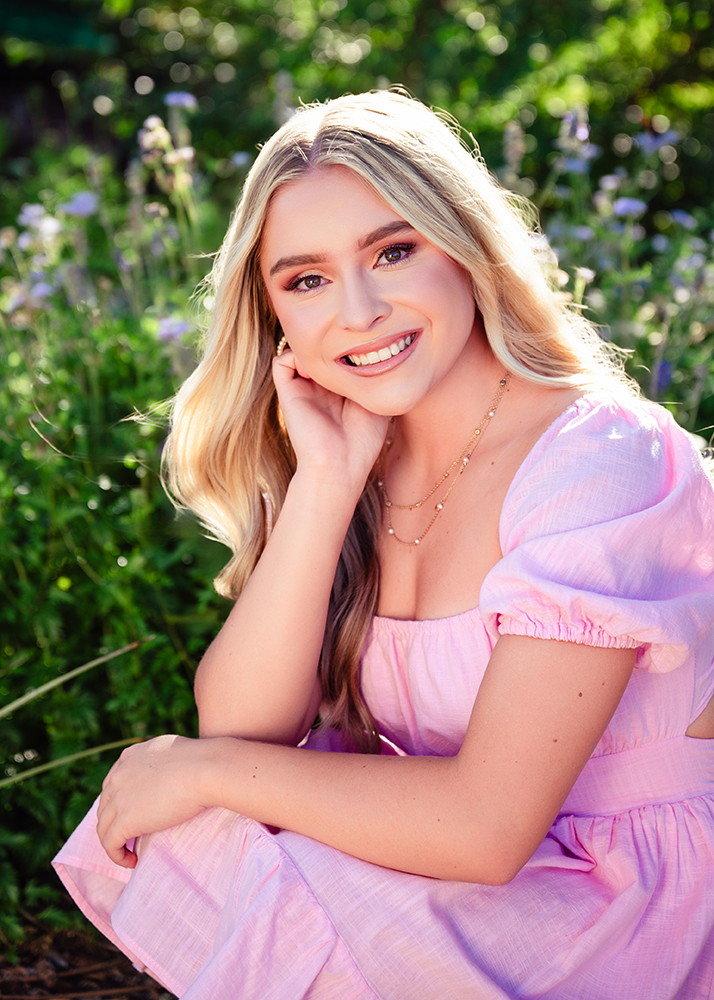 Young woman with long blonde hair smiling warmly, wearing a pink dress, seated outdoors. The background is lush with green plants and purple flowers.