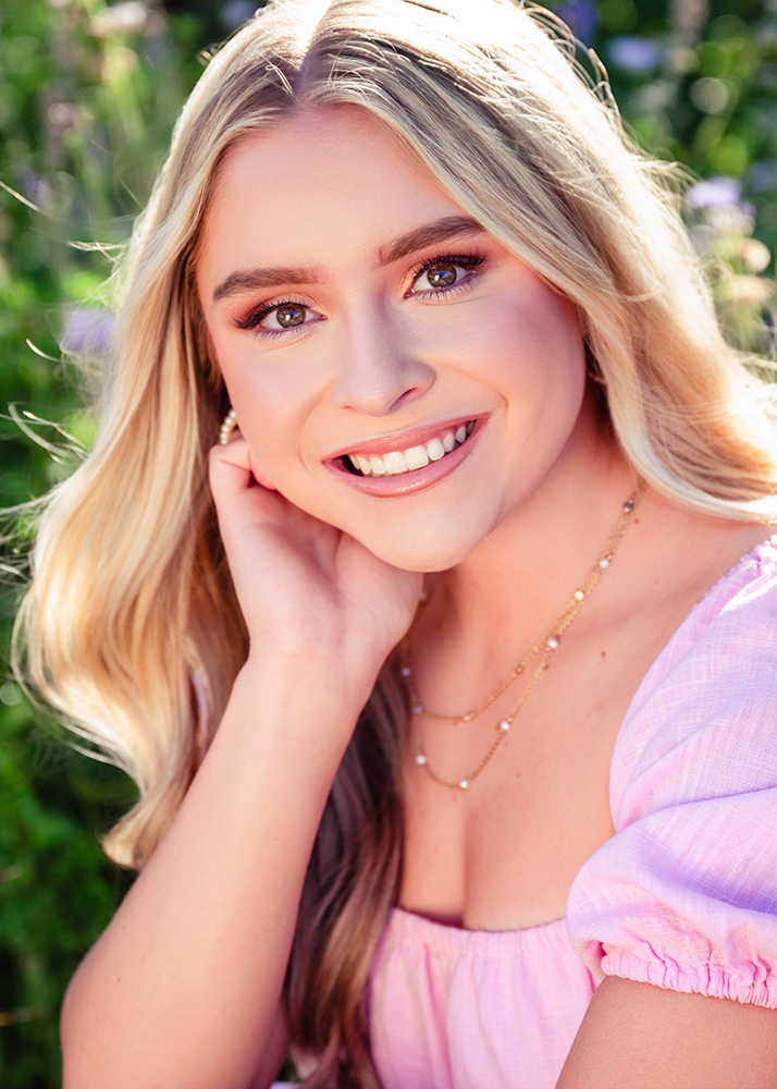 Senior girl smiling warmly, with long blonde hair and wearing a pink top, sits outdoors in a garden. Soft sunlight enhances the joyful and serene mood.