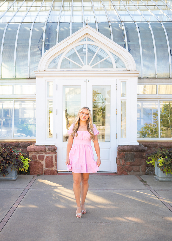 Senior girl in a pink dress stands smiling in front of a glass conservatory with white framing, surrounded by potted plants. Bright, cheerful atmosphere.