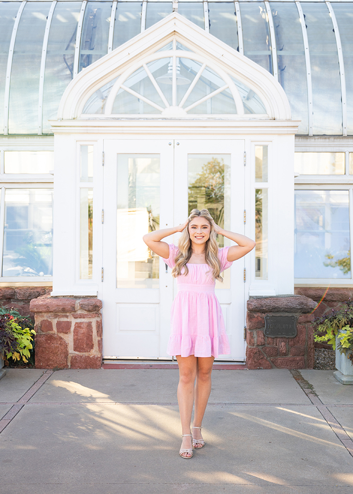 High school senior in a pink dress smiles outside a glass conservatory, standing with hands on her head. Bright sunlight casts soft shadows, creating a cheerful atmosphere.