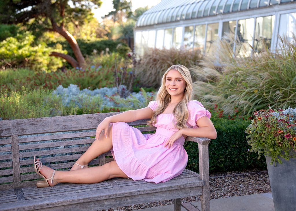 Grace poses for senior photos on a wooden bench at Will Rogers Park in a pink dress in a garden, smiling warmly. The scene is lush with greenery and flowers, conveying a serene, cheerful tone.