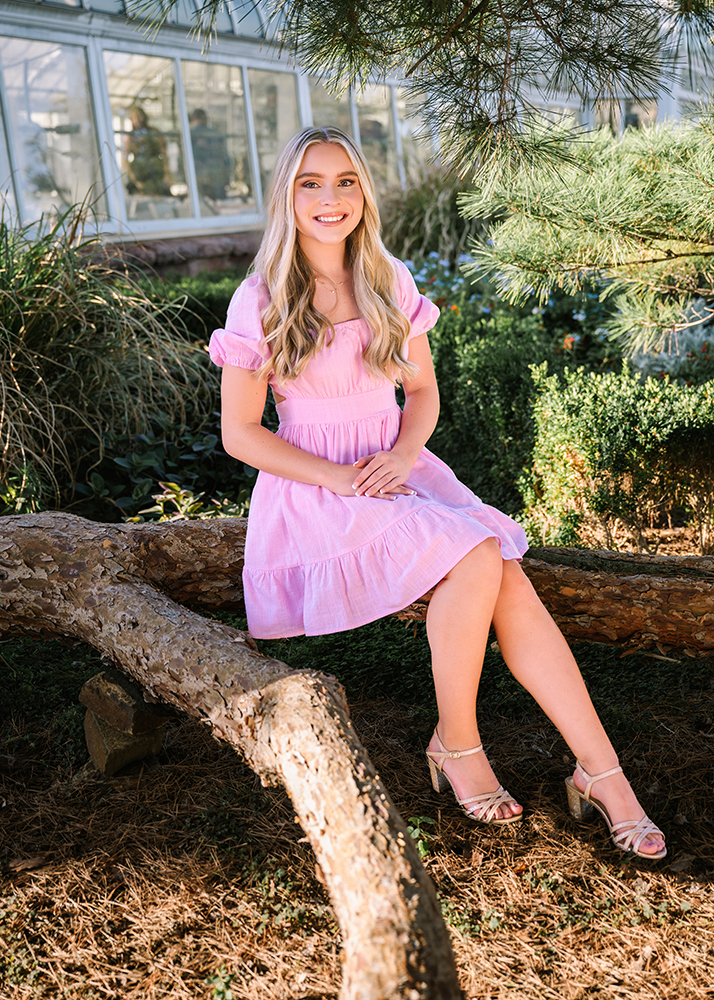 Senior girl with long blonde hair in a pink dress sits on a large curved tree branch. Background features greenery and a glass greenhouse, conveying a serene mood.