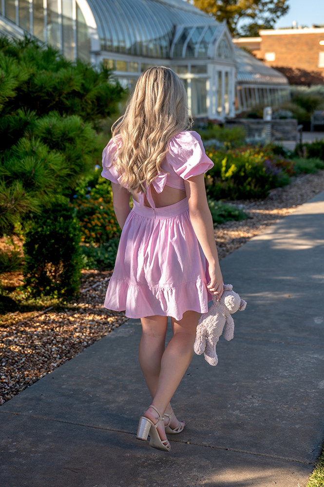 High school senior with long blonde hair walks on a sunlit path, wearing a pink dress and holding her childhood stuffed bear. Greenery and glass buildings surround her.