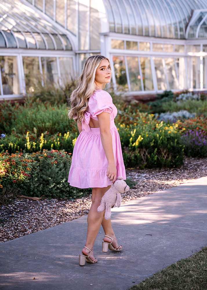 Senior girl in a pink dress with long blonde hair walks on a garden path, holding a plush toy. She gazes back, evoking nostalgia and warmth.