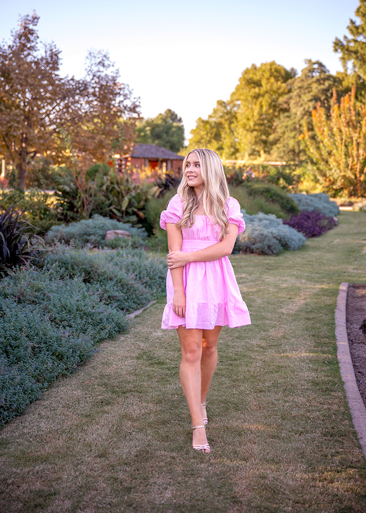 Grace wears a pink dress while she strolls through the sunlit Will Rogers Park garden in OKC, surrounded by lush greenery and trees. She smiles gently, evoking a serene and joyful mood.
