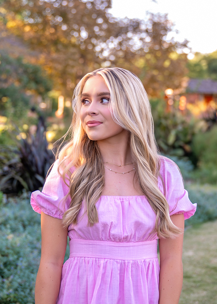 Senior girl in a pink dress stands outdoors, looking to the side. She has long blonde hair and a serene expression. Lush garden background with soft lighting.