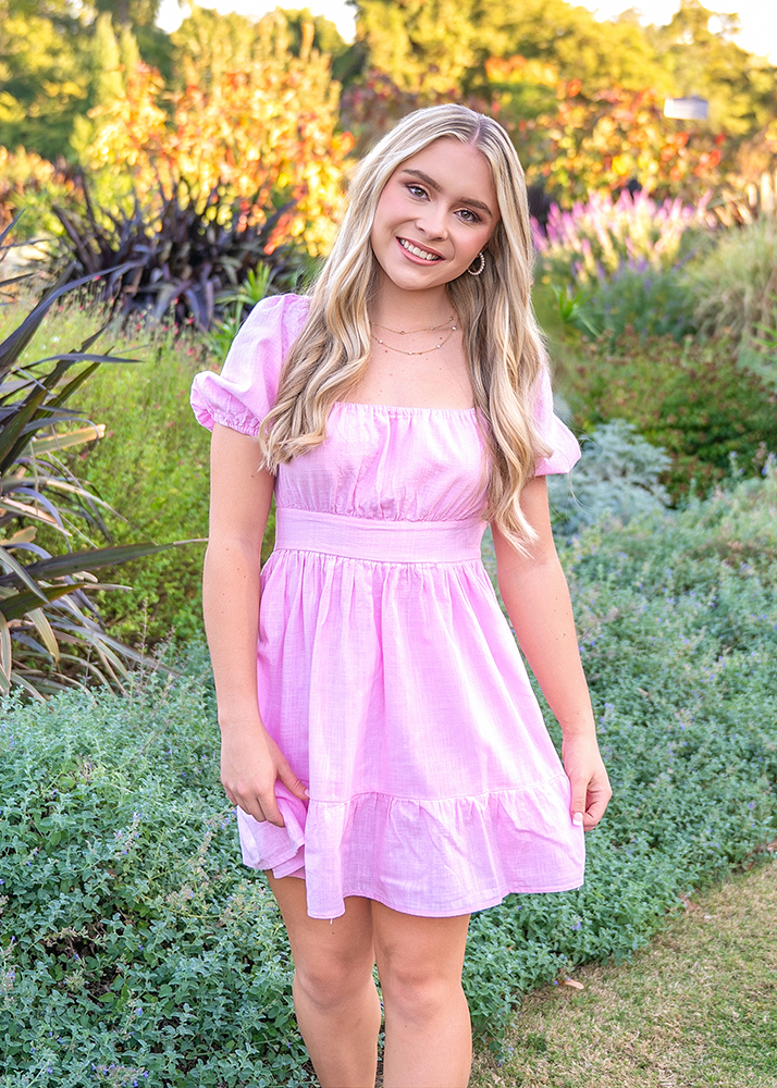Smiling high school senior in pink dress stands in a lush garden. She has long, wavy blonde hair. The sunny setting feels vibrant and cheerful.