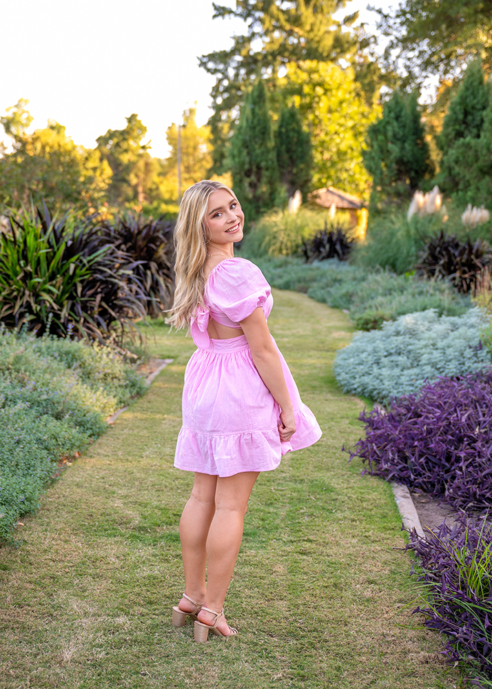 Grace posing for a senior photo in a pink dress on a garden path in Will Rogers Gardens, smiling back at the camera. The setting is lush and green, creating a cheerful and serene atmosphere.
