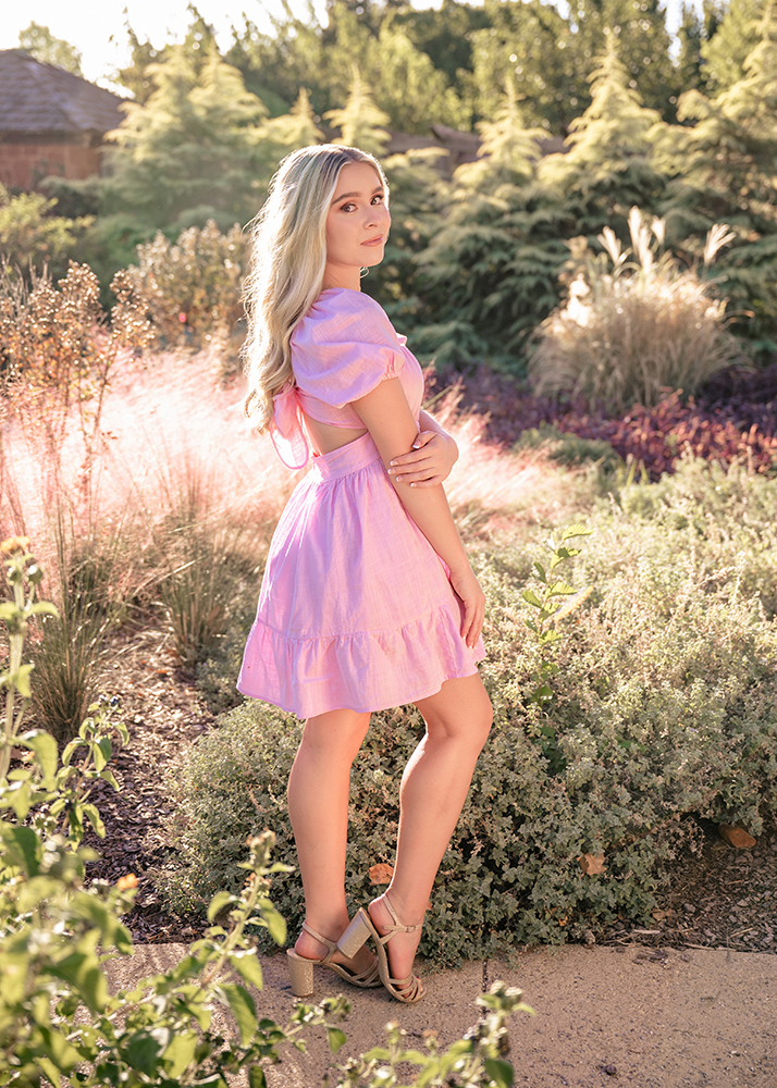 Senior girl in a pink dress stands in a sunlit garden, surrounded by lush greenery. Her expression is serene, exuding a peaceful, gentle vibe.