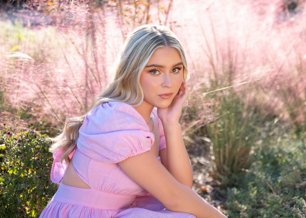 A high school senior in a pink dress sits pensively in a field of soft pink grasses. Sunlight creates a warm, serene glow around her.