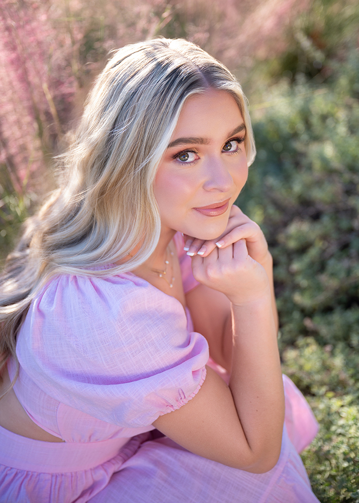 Senior girl with long blonde hair and a pink dress sits in a field of soft pink flowers, smiling gently. The scene is warm and serene.