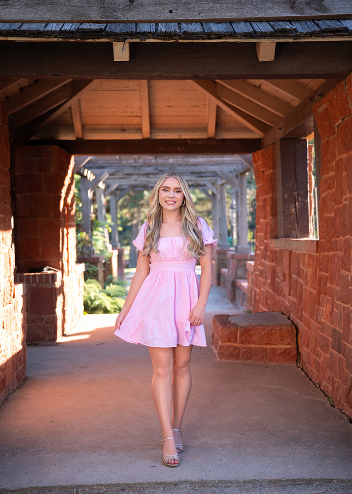 Senior girl in a pink dress stands under a wooden pergola with red brick walls, smiling gently. Sunlight filters through, creating a serene atmosphere.
