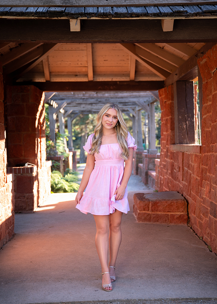 High school senior in a light pink dress walks under a wooden pergola, surrounded by red brick walls and greenery, smiling warmly at the camera.