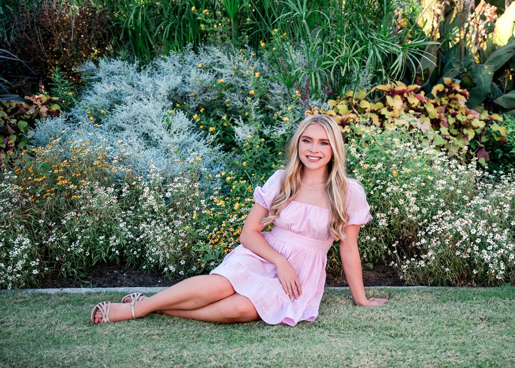 A senior girl in a pink dress sits on grass beside colorful flowers and lush greenery, smiling warmly. The setting is peaceful and vibrant.