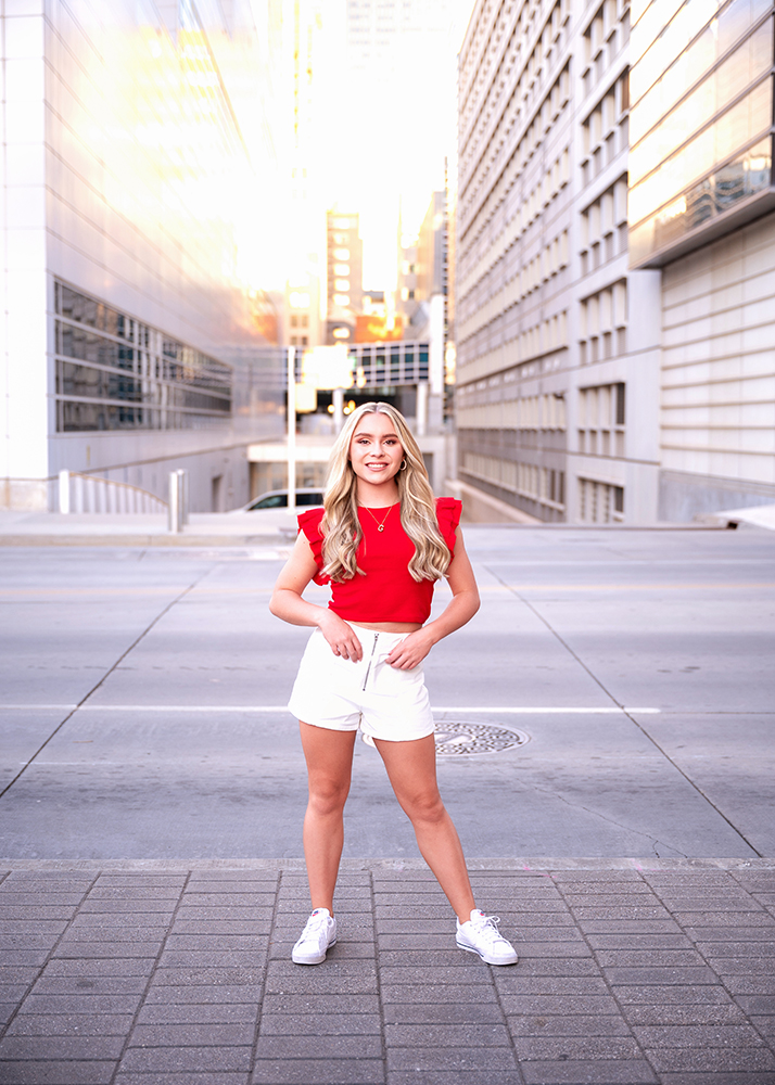 Senior girl with long blonde hair wearing a red top and white shorts stands confidently on an Oklahoma City street, surrounded by tall buildings.