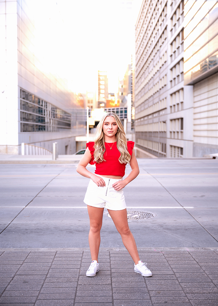 High school senior stands confidently in an urban setting, flanked by modern buildings. She wears a red top, white shorts, and sneakers, exuding a vibrant and poised energy.