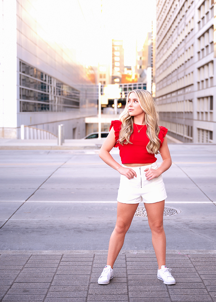 Senior girl in a red top and white shorts stands confidently on a city street, surrounded by tall buildings. The setting sun casts a warm glow.