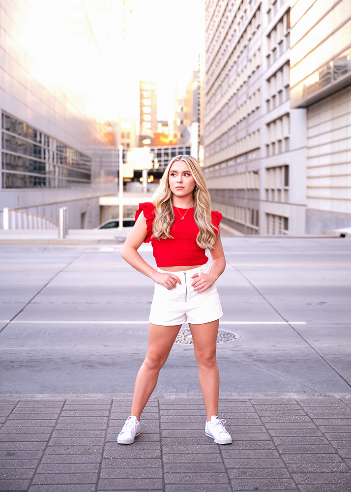 Senior girl with long blonde hair stands confidently on an urban street in a red top and white shorts. She exudes confidence and focus.