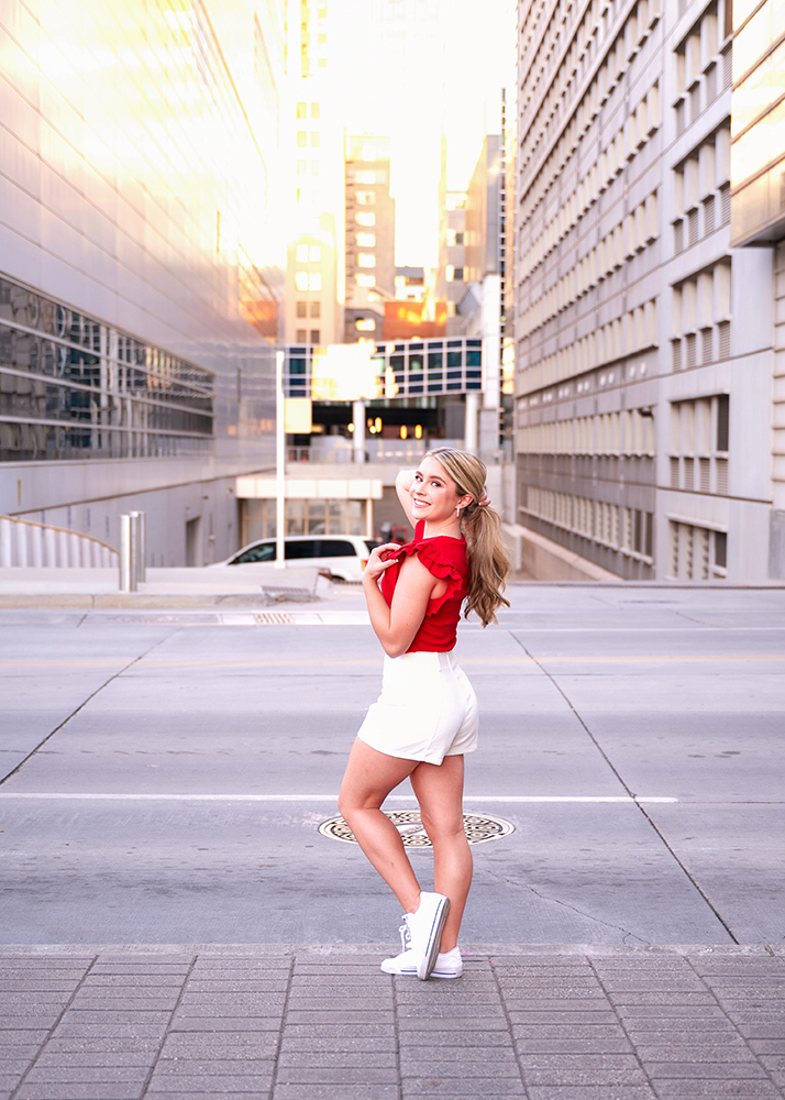 High school senior in a red top and white shorts poses confidently on a city street, surrounded by tall buildings and warm sunlight, conveying a cheerful mood.